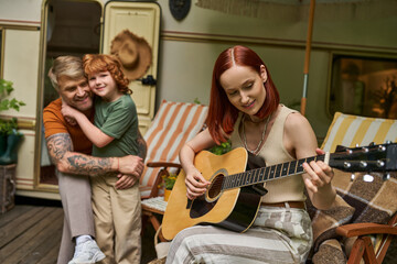 smiling woman playing acoustic guitar near tattooed husband embracing son next to trailer home