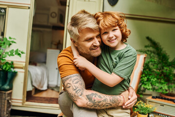 happy tattooed man embracing adorable redhead son near modern trailer home, bonding moments