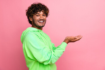 Side photo of cheerful young indian guy wear trendy green hoodie hold two hands asking for charity donate isolated on pink color background