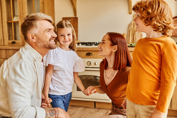 smiling parents with carefree kids holding hands while having fun in kitchen, emotional connection