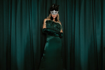 Elegant young woman in green feather dress and crystal carnival mask on dark velvet background in...