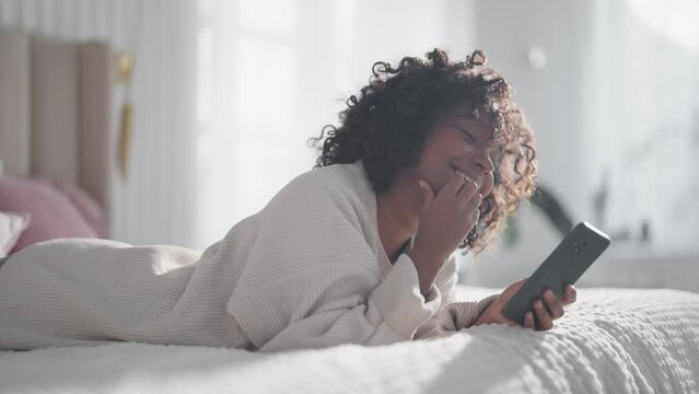 Smiling african american curly woman reading message on smartphone chatting with friend lying in bed at home lazy morning. Positive female enjoying communication in social media looks at phone screen.