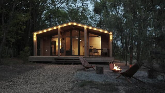 Wooden scandinavian house in the evening, illuminated terrace with warm light from the window, burning fireplace on front of the house. Forest cabin in the middle of a forest in silence, slow motion.