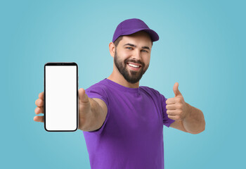 Happy courier holding smartphone with empty screen and showing thumbs up on light blue background