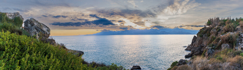 Panorama of the sea with high cliffs on the edges of the frame, sunset behind the distant mountains.