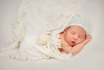Fototapeta na wymiar Newborn baby in a cap with a pompom. The baby is sleeping sweetly. Portrait of a newborn. A newborn baby lies on his tummy and sleeps. Dream. A little boy lies with his cheek on his hand. Son. Baby