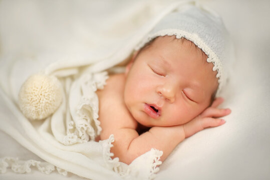 Newborn baby in a cap with a pompom. The baby is sleeping sweetly. Portrait of a newborn. A newborn baby lies on his tummy and sleeps. Dream. A little boy lies with his cheek on his hand. Son. Baby