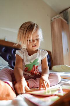 Little girl sits on a bed and reads a book with pictures, running her finger along the lines