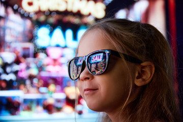 Close-up image of little girl in sunglasses standing on street and looking at shop window. Lights...