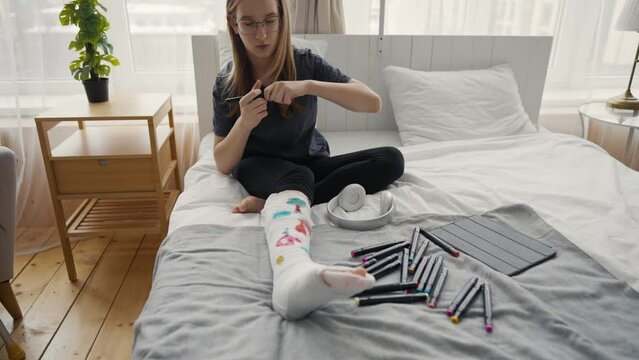 Teen girl painting colorful pictures using felt-tip pens on orthopedic cast on broken leg sitting in bed at home. Boring teenager resting relaxing drawing. Recovery of damaged leg, sick leave concept.