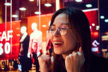 Close-up portrait of smiling young woman with reflection on glasses looking on shop windows. Big sales in shopping mall. Concept of human emotion, shopping, sales, holidays