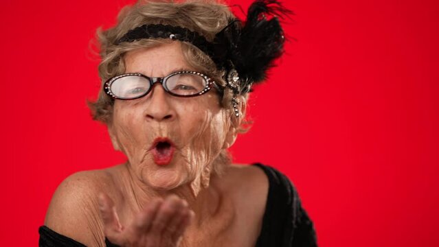 Closeup of fun smiling elderly woman 80s years old walks into frame smiling, blows a kiss and walks off frame, no teeth isolated on red background. Portrait of emotions of love