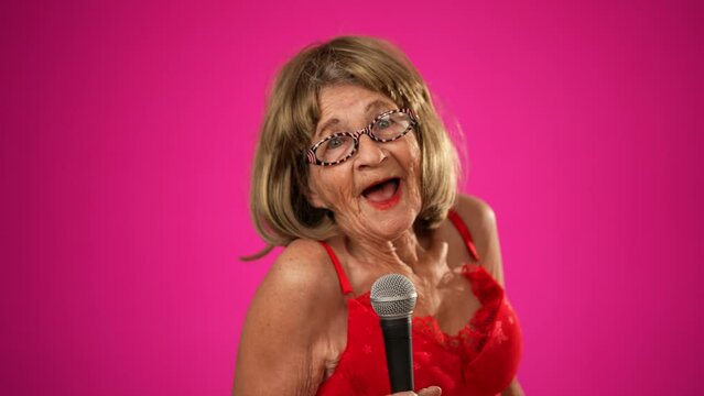 Energetic funny elderly woman with a fashionable look with glasses, singing into a microphone and walking off frame isolated on solid pink background