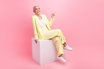 Full size photo of nice woman dressed yellow suit sit on white cube look directing at sale empty space isolated on pink color background