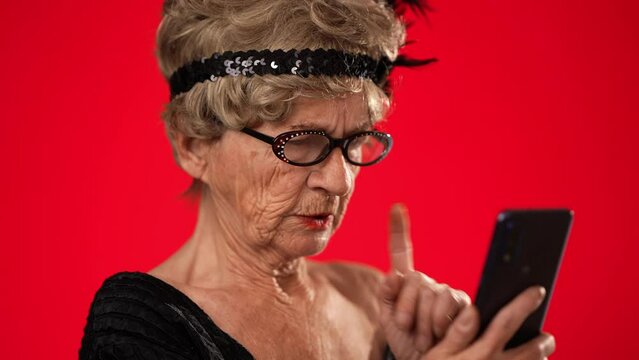 Closeup of funny happy elderly old toothless woman taking selfie using mobile cell phone wearing glasses isolated on solid red background studio portrait. 