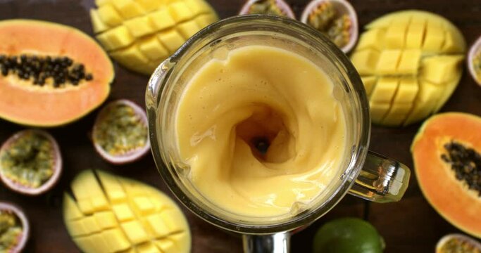 Super slow motion top view of dietetic milkshake smoothie cocktail of organic fresh mango, papaya and passion fruits is being mixed in blender at 1000 fps on wooden table of restaurant.