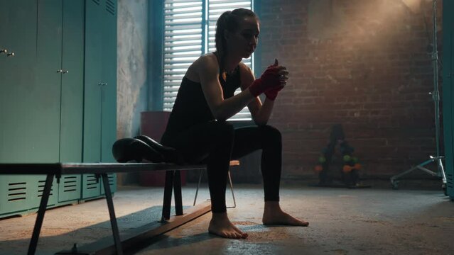 Focused young woman fighter boxer wraps hands wrists with boxing bandage sits on bench in locker room. Girl kickboxer prepares for battle fight. Professional sport, competition, preparation concept.