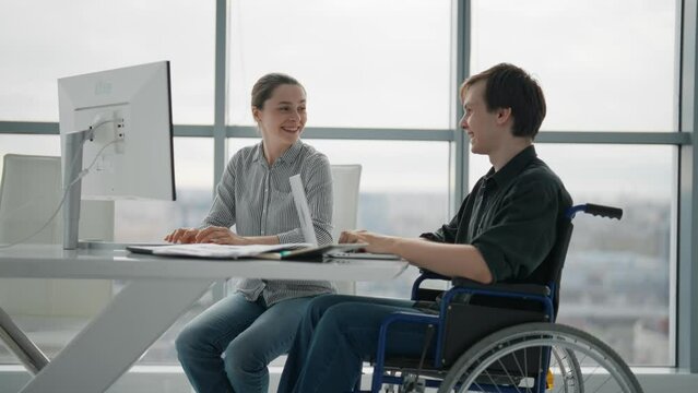 Paralyzed man work with woman in office. Colleagues discussing project together female shows to guy in wheelchair pie chart on paper document. Positive workers discuss analytics data sit at desk.