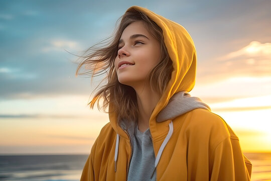 Image generated with AI. Portrait of young woman in hoodie breathing fresh air on the beach at sunrise on a sunny summer day with beautiful warm sky
