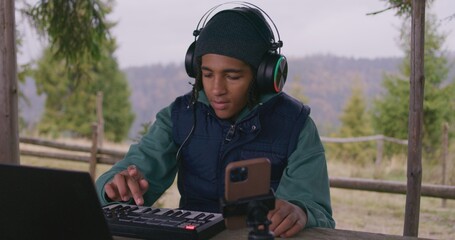 Young musician in headphones plays MIDI controller outdoors. African American teenager creates and...