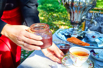 A table with a cup of tea, a samovar and a woman's hand with a jar of jam outdoors on a sunny...