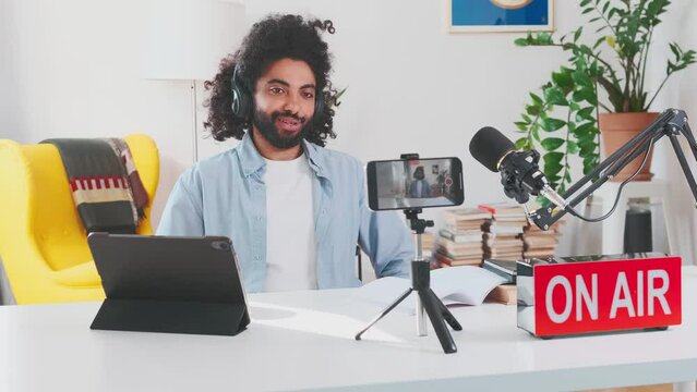 Young enthusiastic Arabian man blogger shoots video on mobile phone camera and applauds announcing results competition on page on social networks sits at table with microphone in home office. On air