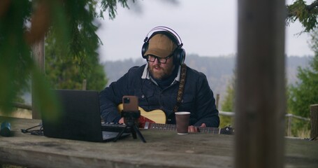 Caucasian man in headphones plays guitar sitting in wooden gazebo in the forest. Male musician composes and records music using laptop and phone on tripod during vacation trip in the mountains.