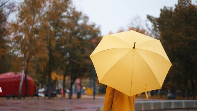 camera tracking shot of woman in yellow raincoat with bright umbrella walks in autumn park at rainy day. wet rain weather, fall season, golden foliage