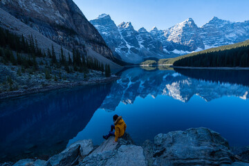 Female tourist carries a backpack, travels, walks, climbs, sits and admires the beauty of the reflection in the water of moraine Lake, Canadian Rocky Mountains - obrazy, fototapety, plakaty