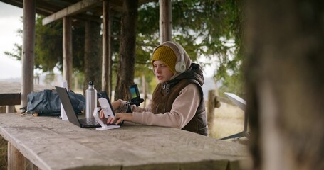 Teenager in headphones uses laptop and phone on tripod to create content for viewers during holidays in mountain forest. Young streamer sits in wooden gazebo and talks into professional microphone.