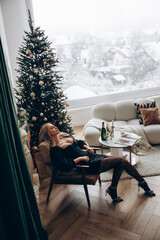 Young pensive woman sitting in armchair in black dress with decollete near Christmas tree and table.
