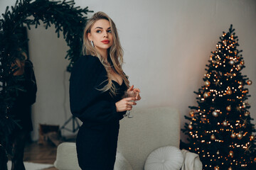 Young pensive blonde woman standing in decollete dress with wine glass in her hands near Christmas tree.