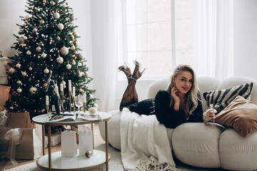 Young blonde woman lying down on sofa in black dress and high heel shoes near decorated Christmas tree and table.