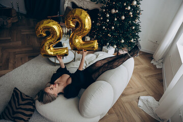 Young woman celebrating her birthday, lying on sofa and holding figures of numbers 22.