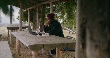 African American business man sits in wooden gazebo, talks at online conference using laptop and phone on tripod. Traveler remotely consults clients during vacation in beautiful forest. Outdoor work.