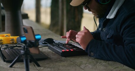 Close up of Caucasian musician sitting at table in gazebo with laptop, phone on tripod and musical...
