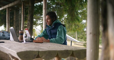 African American young boy sits in wooden gazebo, talks with teacher at video call using laptop and phone on tripod. Tourist remotely studies during holidays in mountain forest. Outdoor study concept.