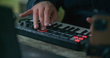 Close up of male musician playing digital electric piano and composing music, sitting outdoors in forest. Caucasian man uses laptop and phone on tripod to record song on his vacation in mountains.