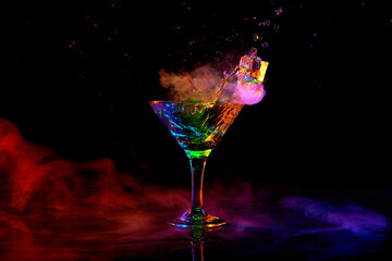 Glass with delicious cocktail with martini against dark background with neon light with smoke. Pouring liquid, splashes. Concept of alcohol drink, nightclub, party, taste, celebration.