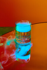 Glass with strong drink, whiskey and smoke on top standing against orange background in neon light. Concept of alcohol drink, nightclub, party, taste, celebration.