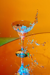 Ice cube falling into glass with martini cocktail against orange background in neon light. Splashes. Concept of alcohol drink, nightclub, party, taste, celebration.