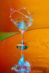 Ice cube falling into glass with martini cocktail against orange background in neon light. Splashes. Concept of alcohol drink, nightclub, party, taste, celebration.