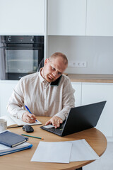 Fototapeta premium a Caucasian man, 35-39 years old, in a light shirt, working at a laptop and talking on a mobile phone, making notes in a notebook, in a light kitchen.a lot of papers and cups of coffee on the table.