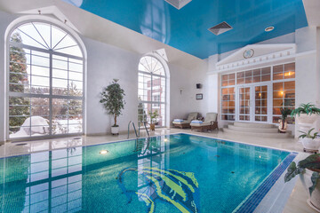 Exclusive interior of a swimming pool in a luxurious mansion. Lounge chairs at the edge .of the pool. Windows with a view of the winter landscape.