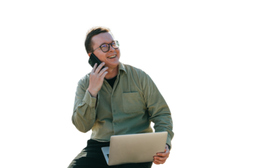 Cheerful young man in glasses and casual sitting against transparent background with laptop talks by phone toothy smiles enjoying conversation with friends. Relocation, freelancer works.