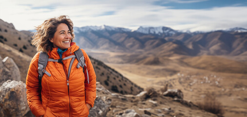 Mature woman in orange jacket hiking traveling in highlands,standing enjoying mountain landscape. - obrazy, fototapety, plakaty