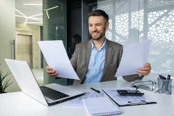 Portrait successful financier accountant at workplace inside office, man looks, checks reports, holds contract papers in hands, businessman in business suit smiling, satisfied achievement results.