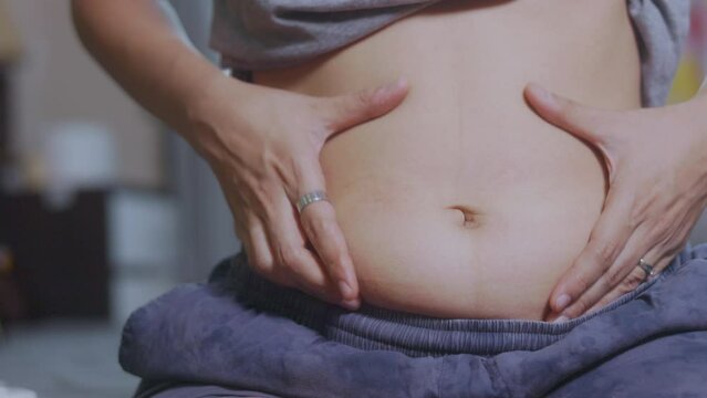 Close-up shot of her hand touching and shaking her fat belly and cellulite of an obese Asian womanin bed. The stretched skin on her abdomen emphasizes her weight gain and concerns about her health. 