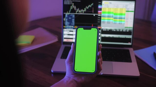 Trader holds smartphone in hands with green screen on chart of movement of cryptocurrency on screen of monitor laptop background. Stock market data statistics using mobile app.