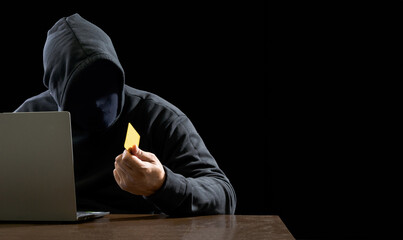 Hacker spy man one person black hoodie sitting on table hand holding credit card look computer...
